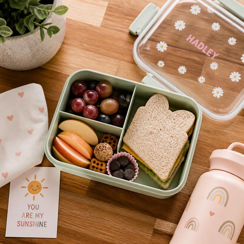 A neatly packed bento-style school lunch box with fruit, a sandwich, and snacks on a wooden kitchen counter